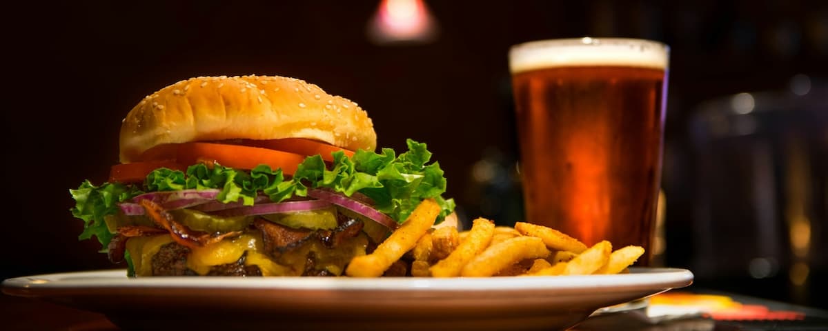 A sesame seed bun burger with lettuce, tomato, onion, pickles, cheese, and a side of fries, paired with a glass of beer.