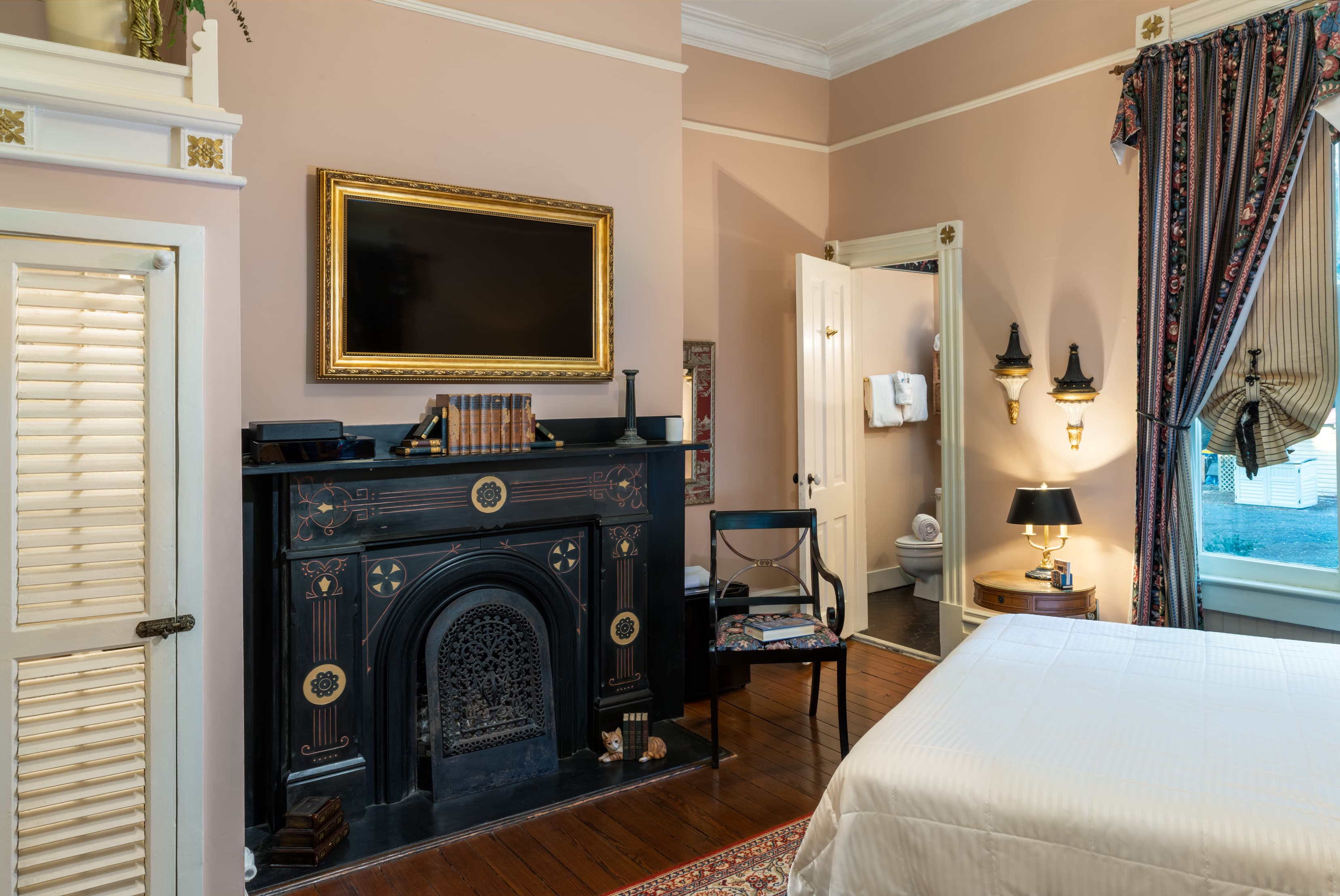 Elegant guest room featuring a decorative black fireplace, high ceilings, hardwood floors, and a cozy queen bed with historic Victorian-style décor.