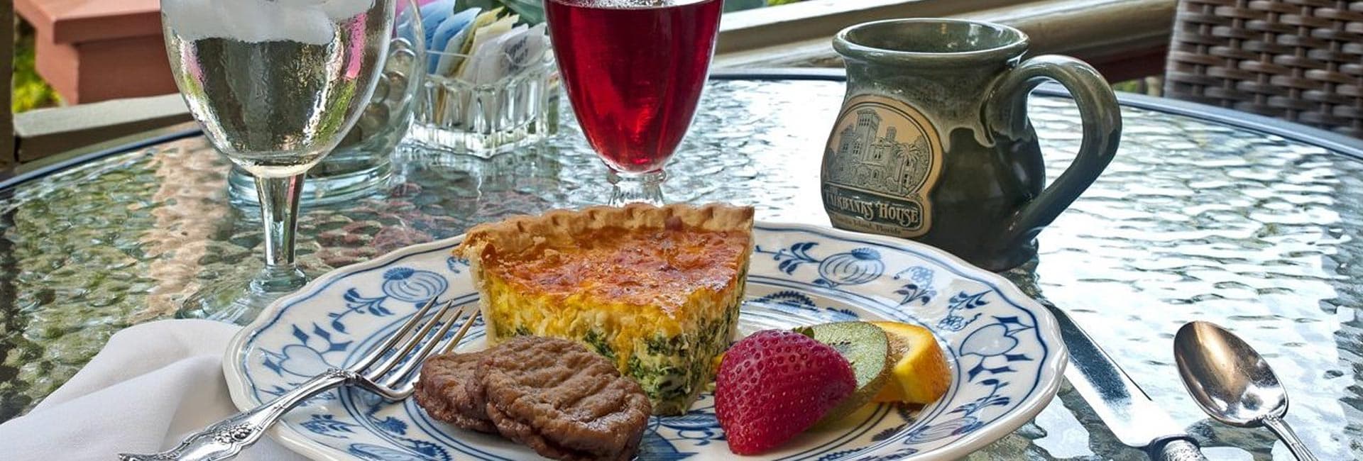 A plate with a slice of quiche, two sausage patties, a strawberry, and a lime wedge, accompanied by a cup of coffee and a glass of iced drink on a table.