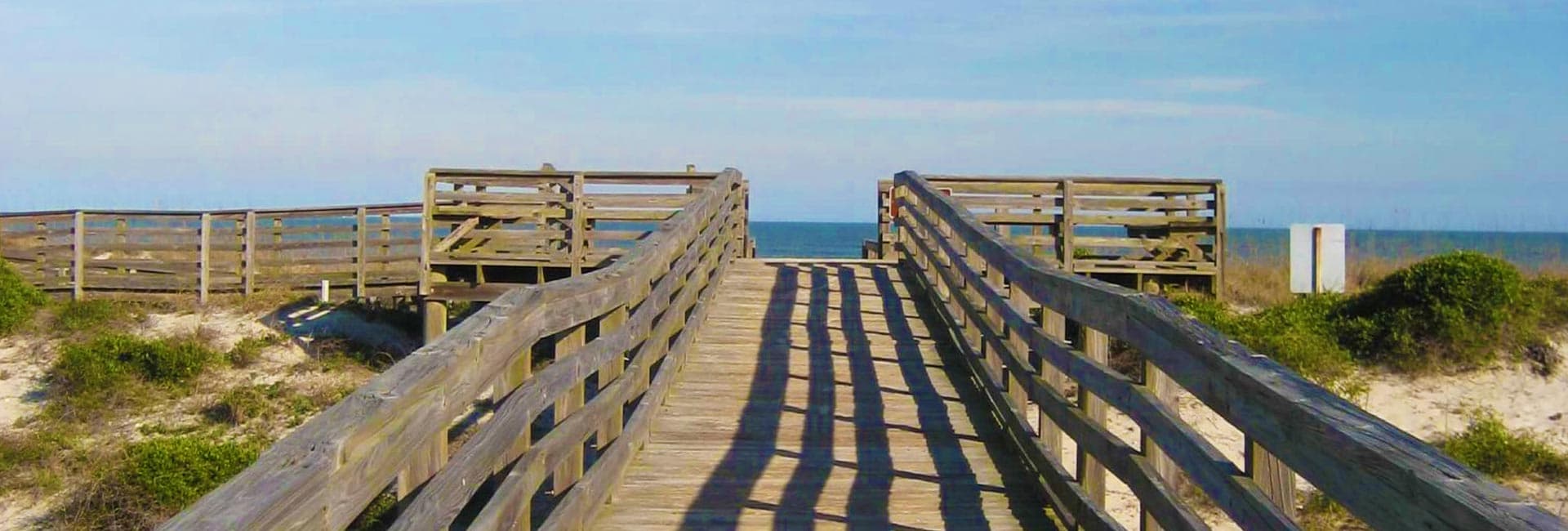 A wooden boardwalk leads to a sandy beach and the ocean under a clear blue sky.