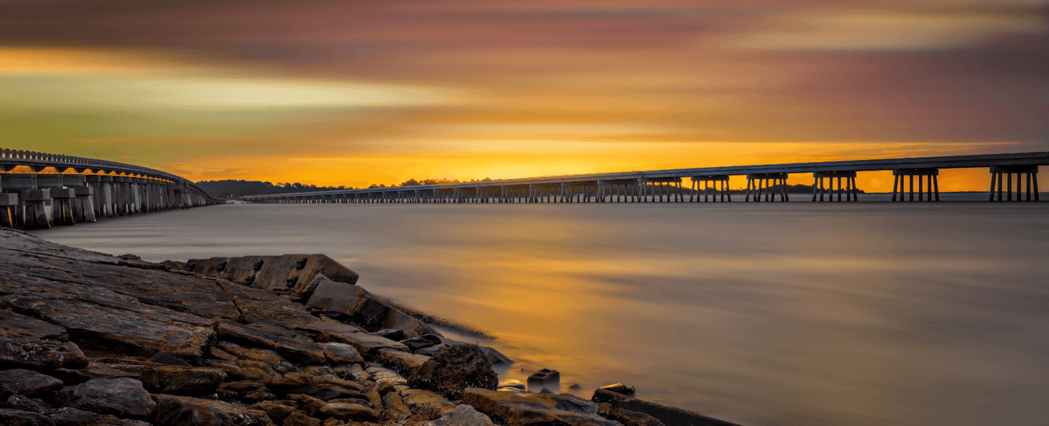 A scenic view of a long bridge stretching over calm water at sunset, with colorful clouds in the sky.