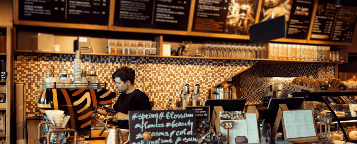 A barista prepares drinks at a stylish coffee shop with a modern decor and baked goods display.