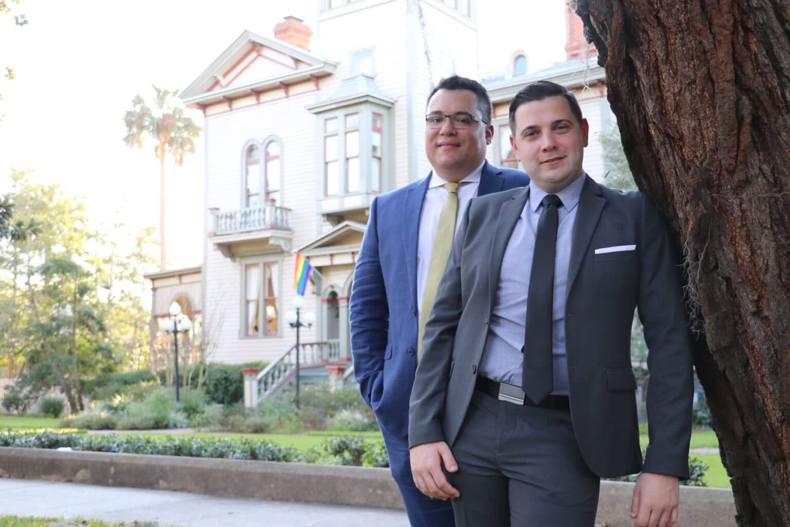 Two men in suits pose in front of a historic house with a rainbow flag.