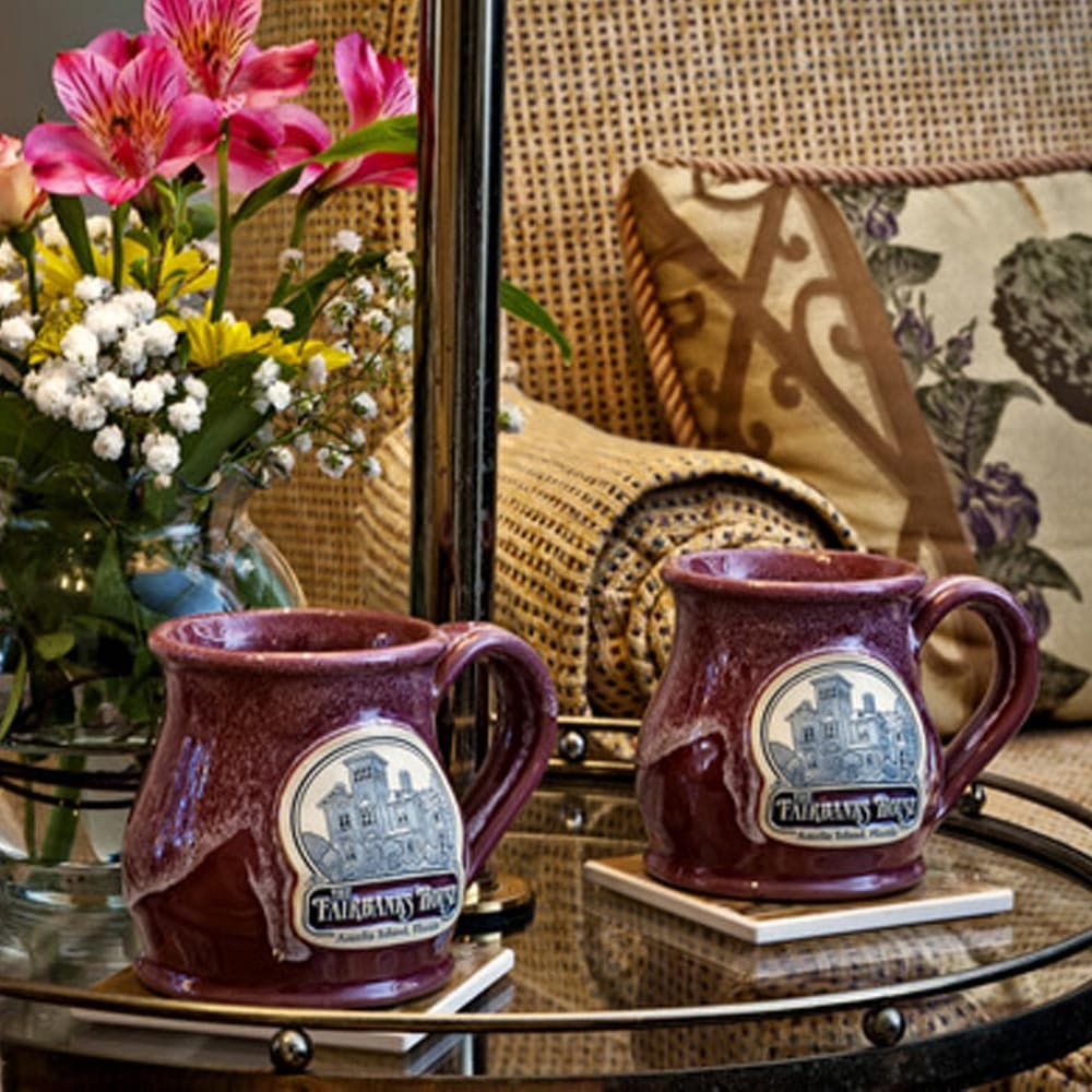 Two purple mugs with a logo sit on coasters beside a vase of flowers on a glass table.