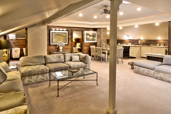 Very large living room with sloped ceilings. Large sectional couch encircles the main area. Dining table and chairs for 6, along with Kitchen and entrance to one bedroom in background.