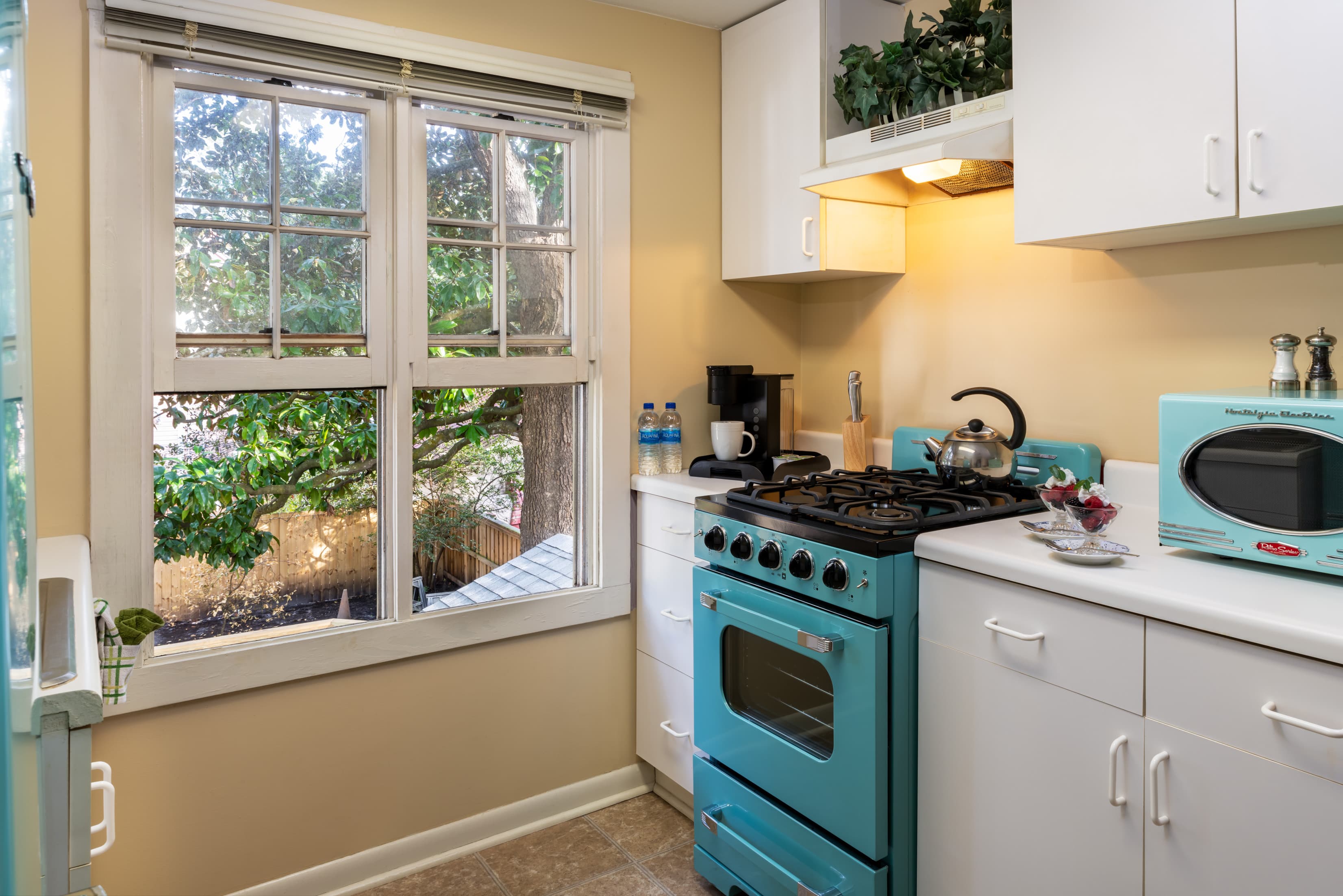 Bright cottage kitchen with retro-style turquoise stove and toaster, white cabinetry, coffee station, and large window overlooking the grounds.