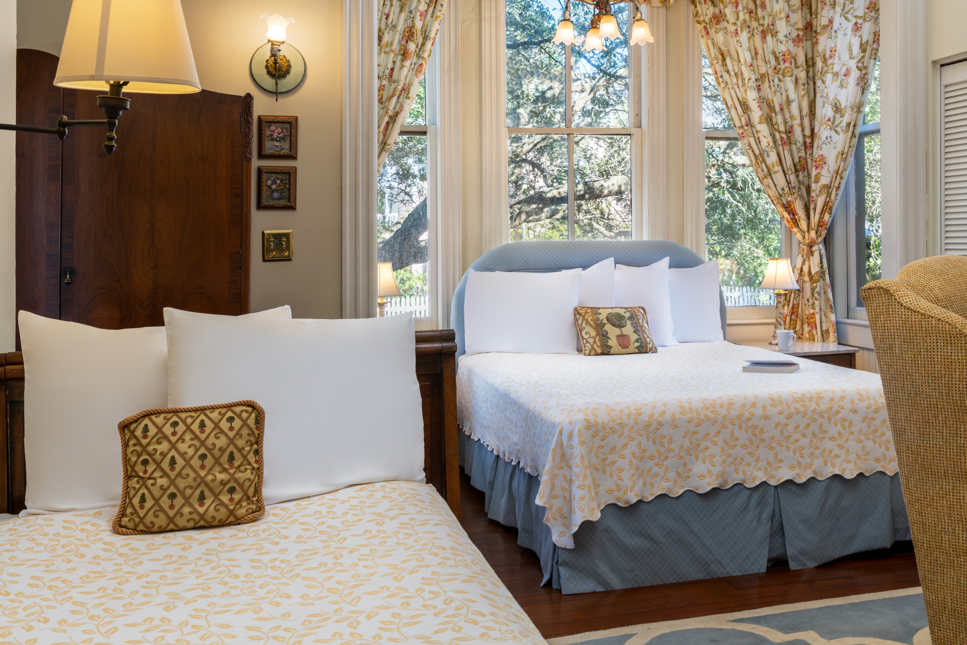 Bright and inviting guest room at featuring two beds with elegant patterned bedding, large windows with floral drapes, hardwood floors, and abundant natural light overlooking oak trees outside.