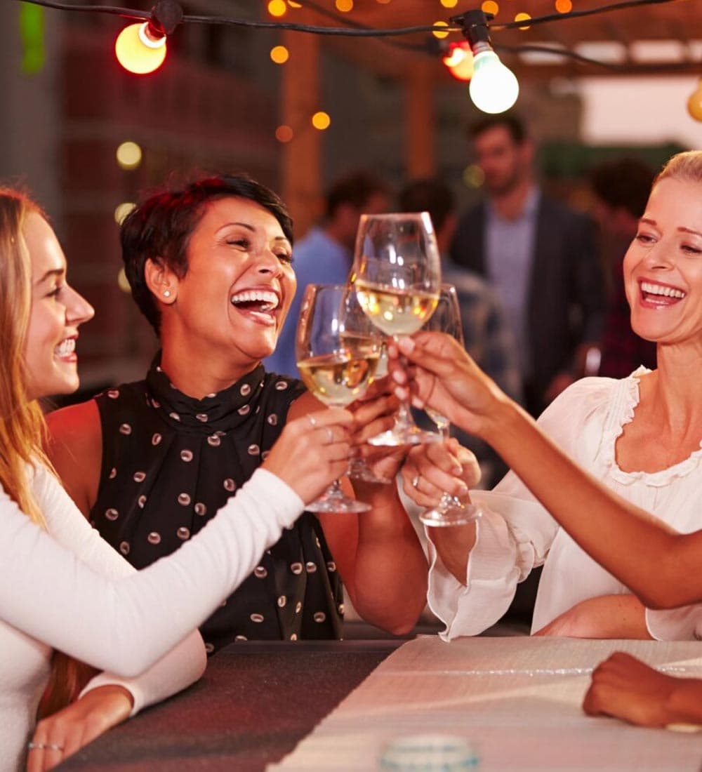 Three women joyfully clink glasses of white wine in a festive outdoor setting.