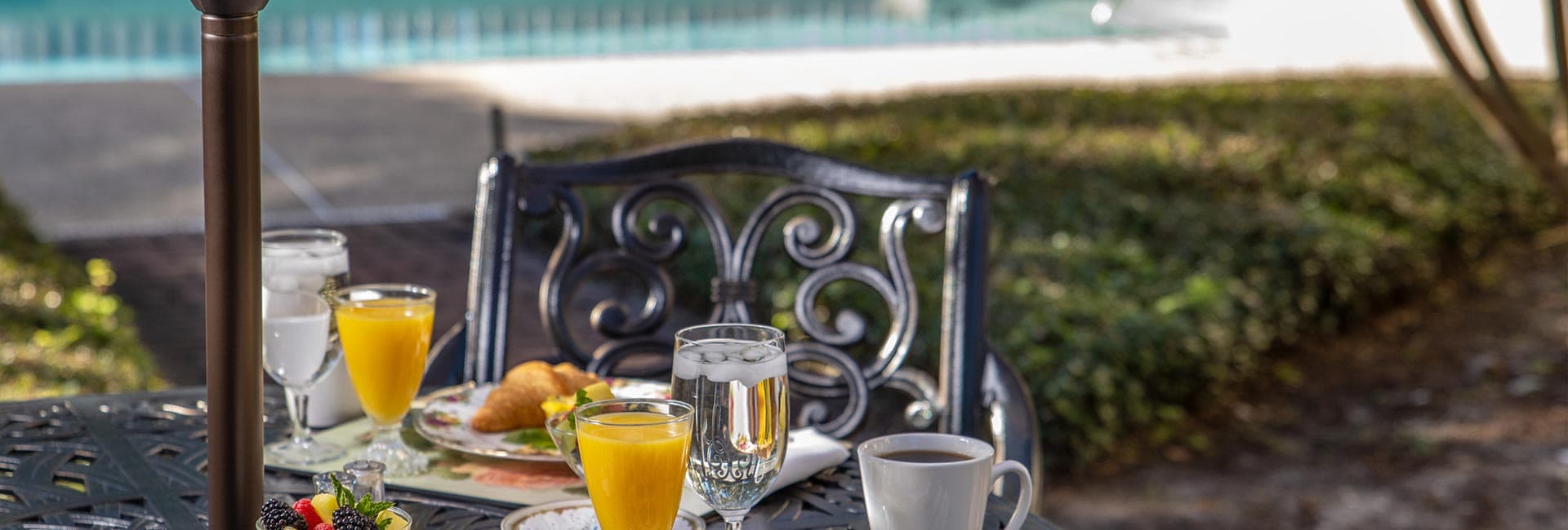A beautifully set outdoor table with breakfast items, including juice, coffee, and pastries, beside a pool.
