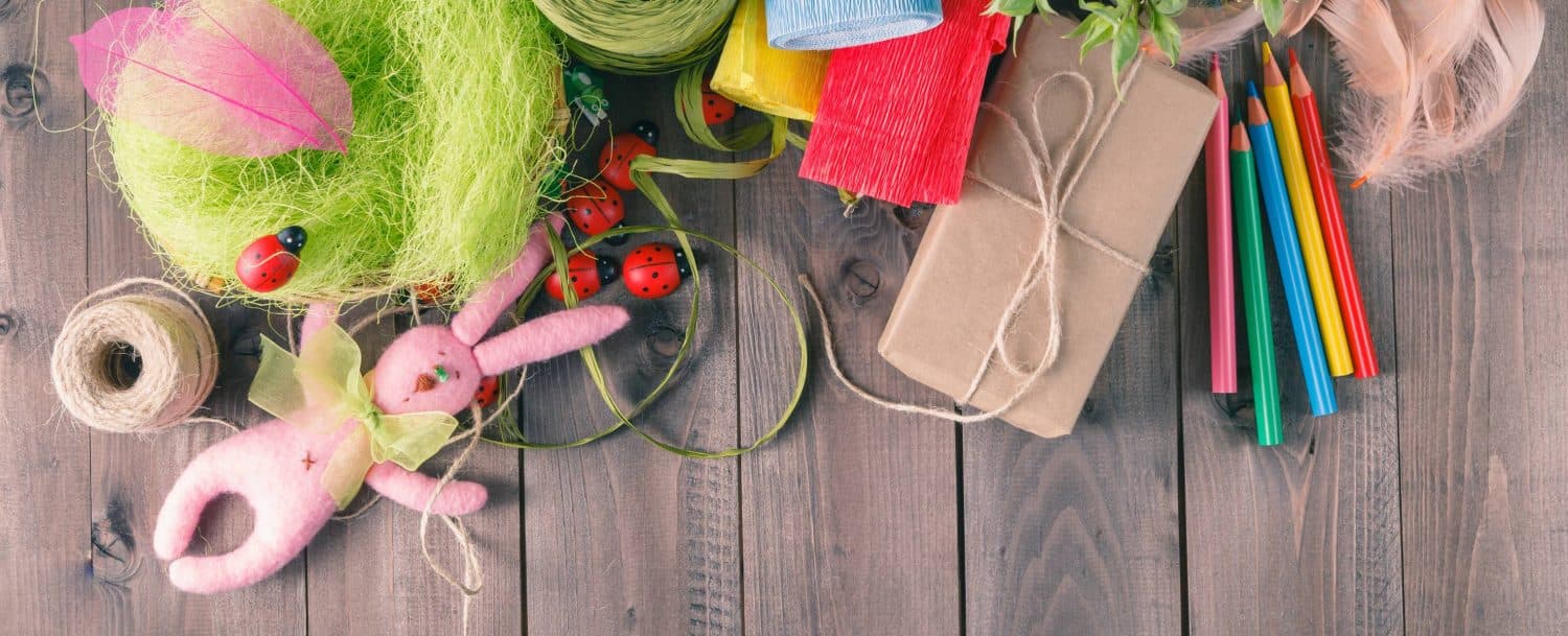 A colorful assortment of crafting materials, including yarn, scissors, a gift box, colored pencils, and a pink bunny toy, arranged on a wooden surface.