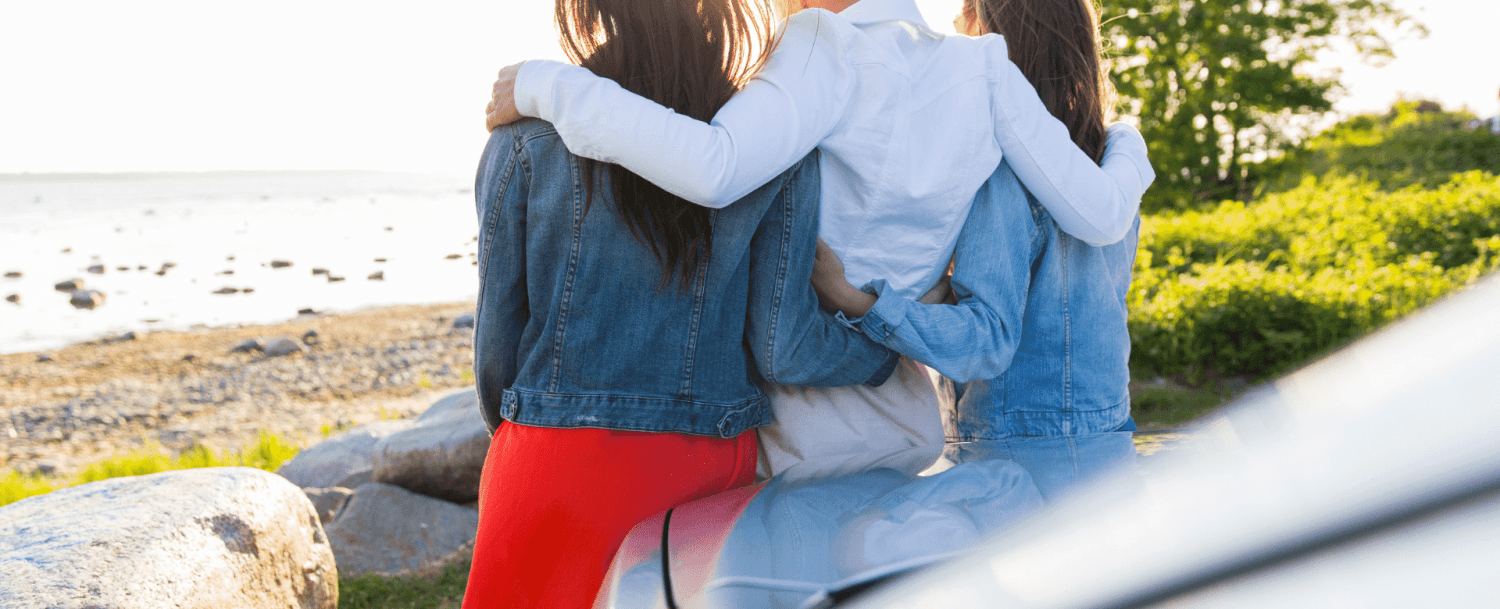 Three women in casual outfits embrace while sitting on a rocky shore, facing a scenic waterfront. Three women in casual outfits embrace while sitting on a rocky shore, facing a scenic waterfront.