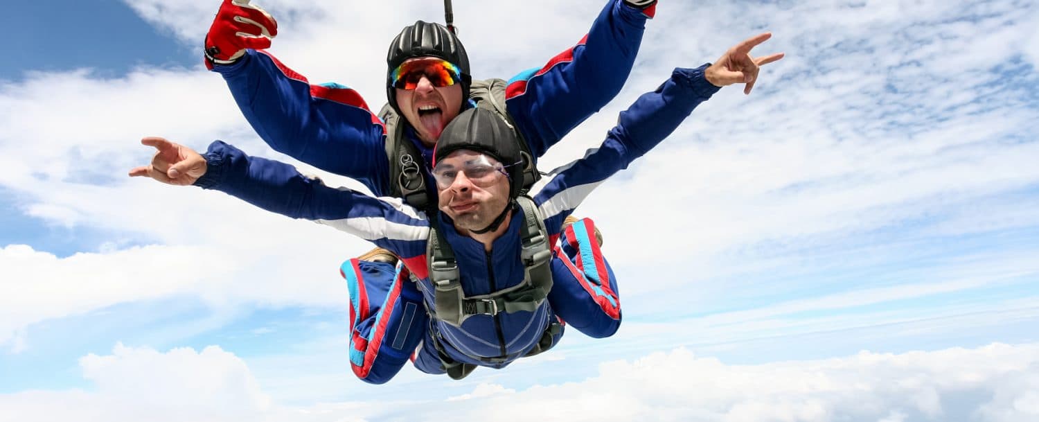 Two skydivers in jumpsuits free-fall through a cloudy sky, one in front making a playful expression.