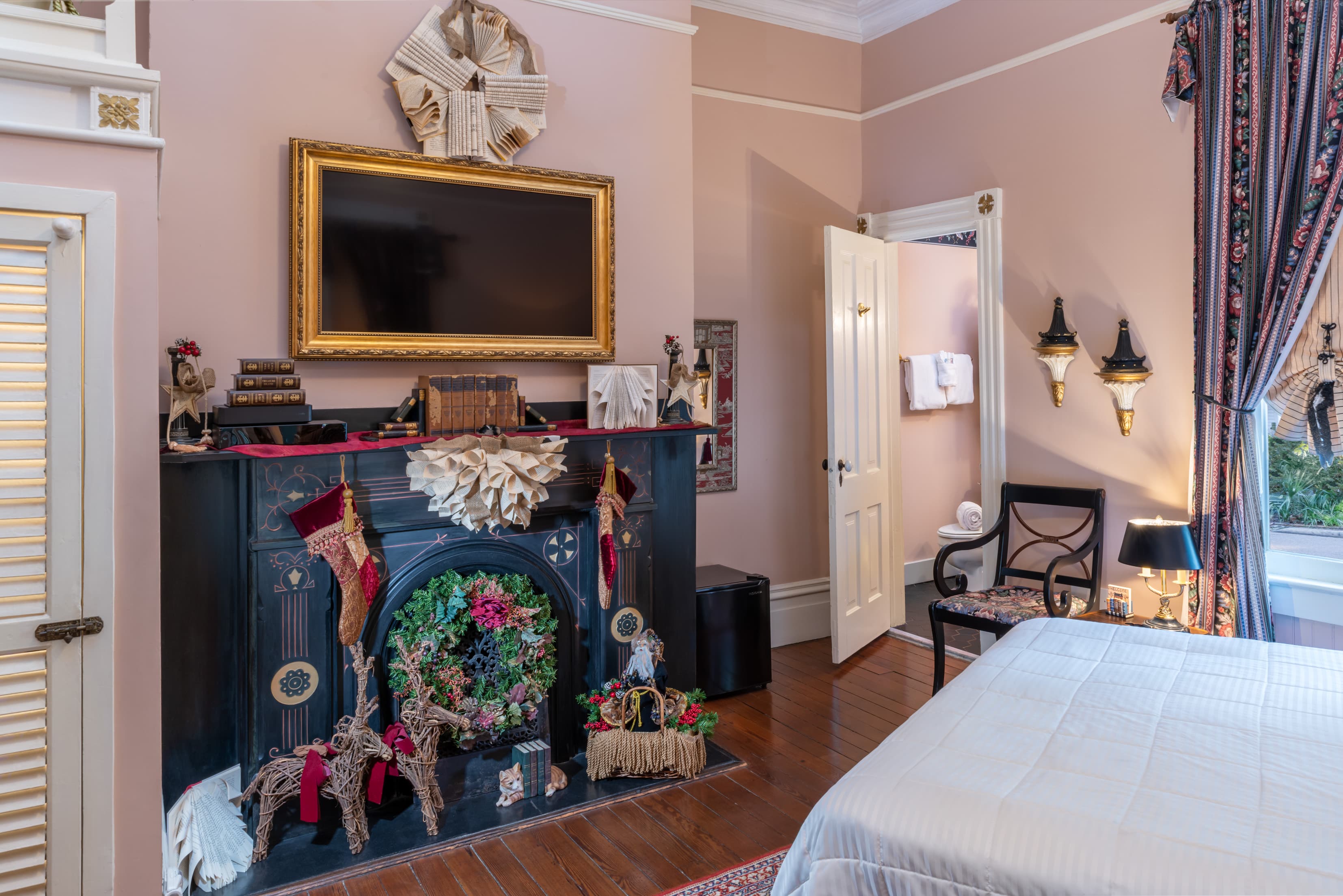 Elegant guest room at featuring blush pink walls, a decorative black fireplace with festive holiday décor, a gold-framed television above the mantel, hardwood floors, patterned drapes, and a cozy bed beside a sitting chair and soft bedside lighting.