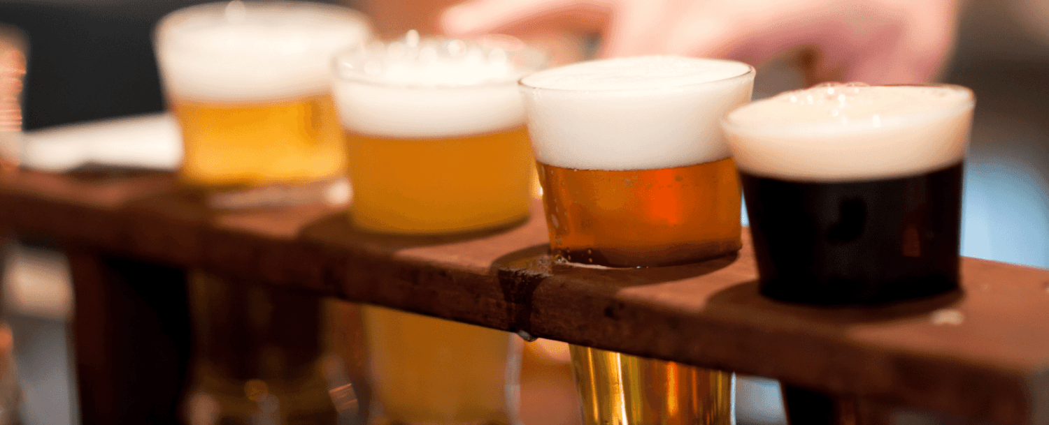 A flight of four small glasses of various beers on a wooden paddle.