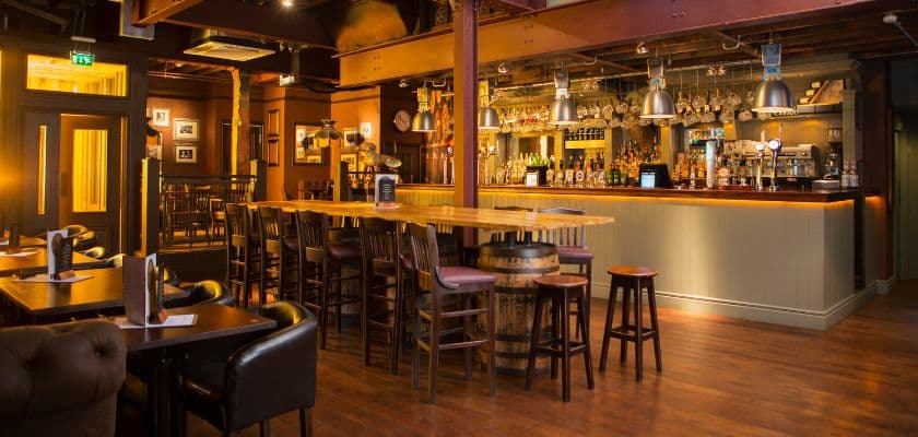 A warm, inviting pub interior featuring a long wooden bar, high stools, and ambient lighting.