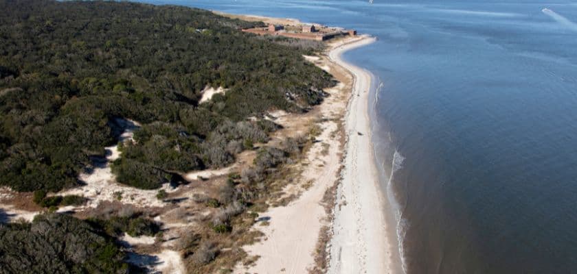 Aerial view of a sandy beach bordered by greenery along a calm shoreline.
