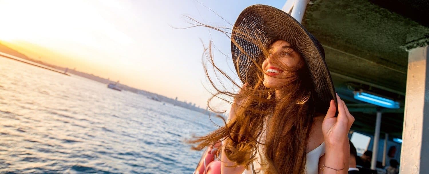 A young woman in a wide-brimmed hat smiles as her hair blows in the wind, with a sunset over the water in the background. A young woman in a wide-brimmed hat smiles as her hair blows in the wind, with a sunset over the water in the background.
