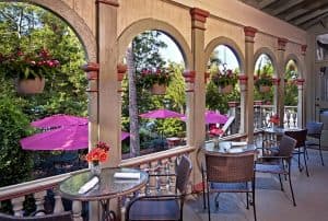 A charming outdoor dining area with arched columns, tables, and purple umbrellas surrounded by greenery.