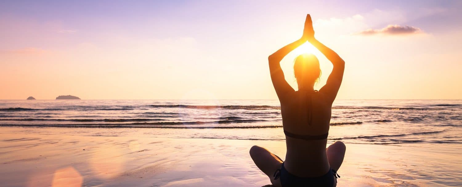 A person sits in a yoga pose on the beach, silhouetted against a sunrise over the ocean.