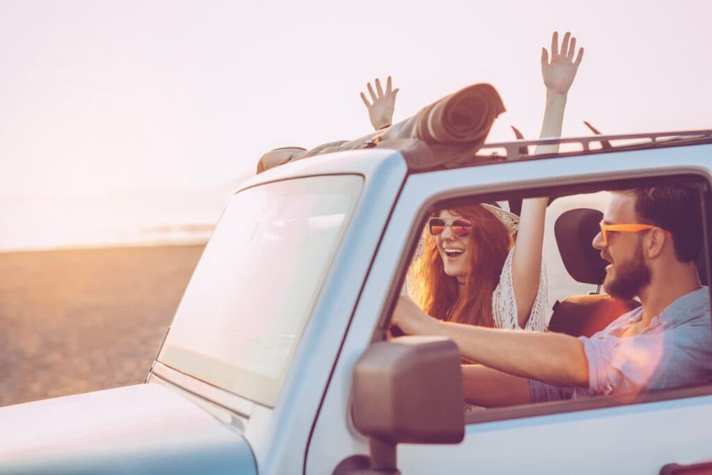 A joyful couple drives with their arms raised in a convertible, enjoying a sunset at the beach.