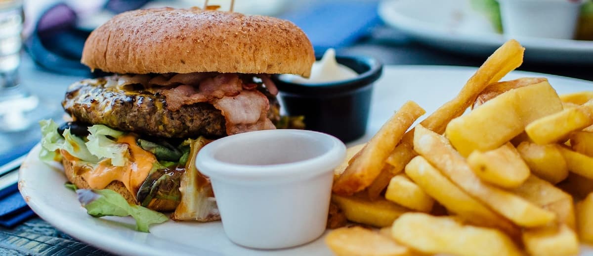 A juicy hamburger with bacon, lettuce, and cheese, served with golden fries and dipping sauce on a plate.