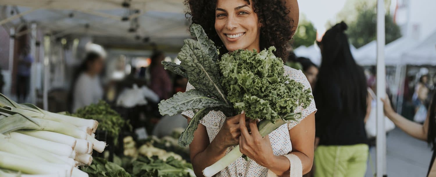 A smiling woman with curly hair holds fresh kale and other vegetables at a market.