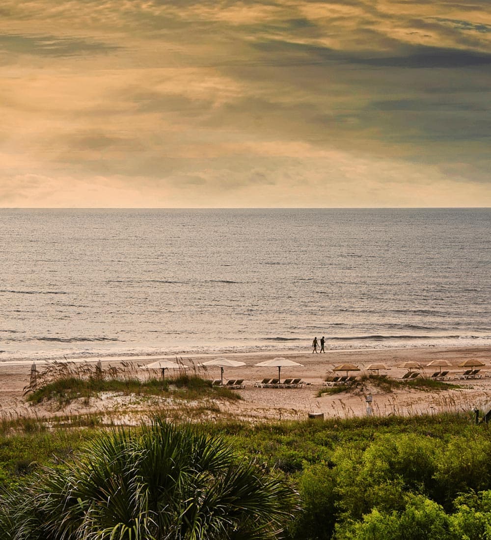 A couple walks along a sandy beach under a colorful sky.