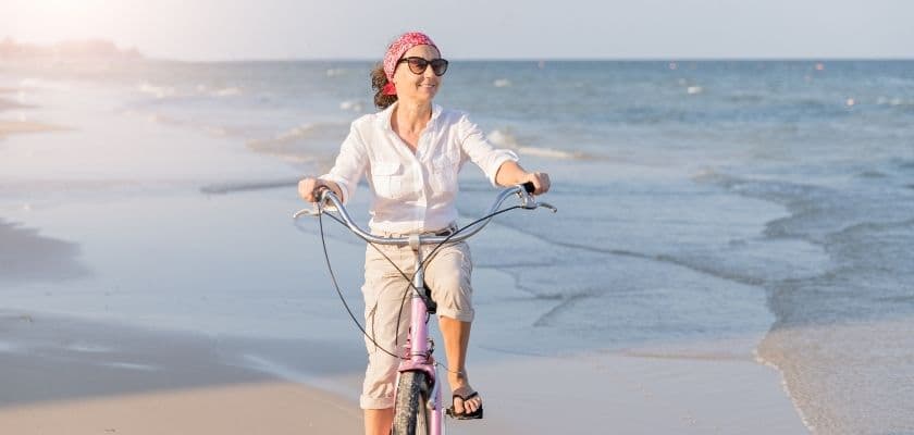 A woman with sunglasses and a headband rides a bicycle along a beach.