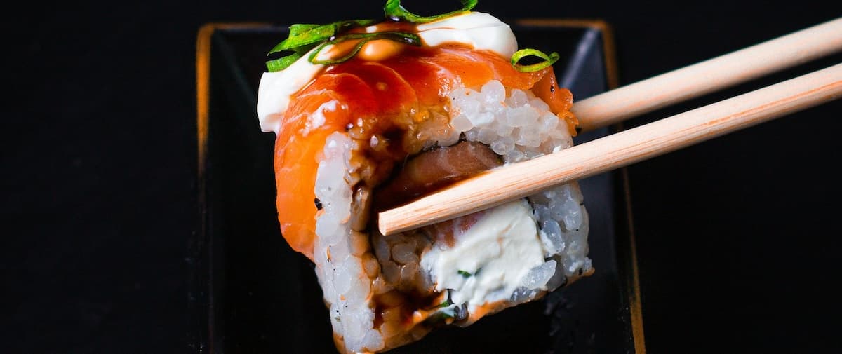 Close-up of a sushi roll with salmon, cream cheese, and green onions, held with chopsticks.