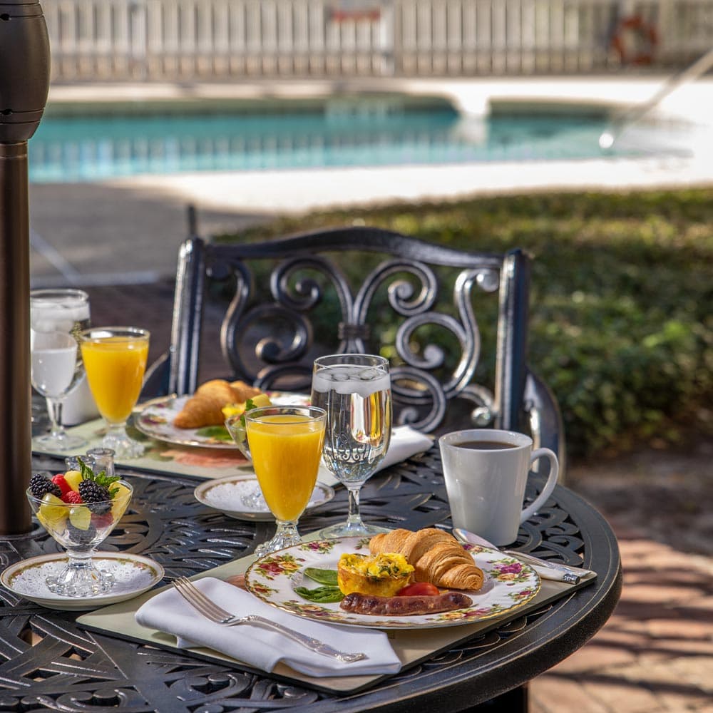 A beautifully arranged outdoor breakfast table with a pool view, featuring fruit, pastries, drinks, and neatly folded napkins.