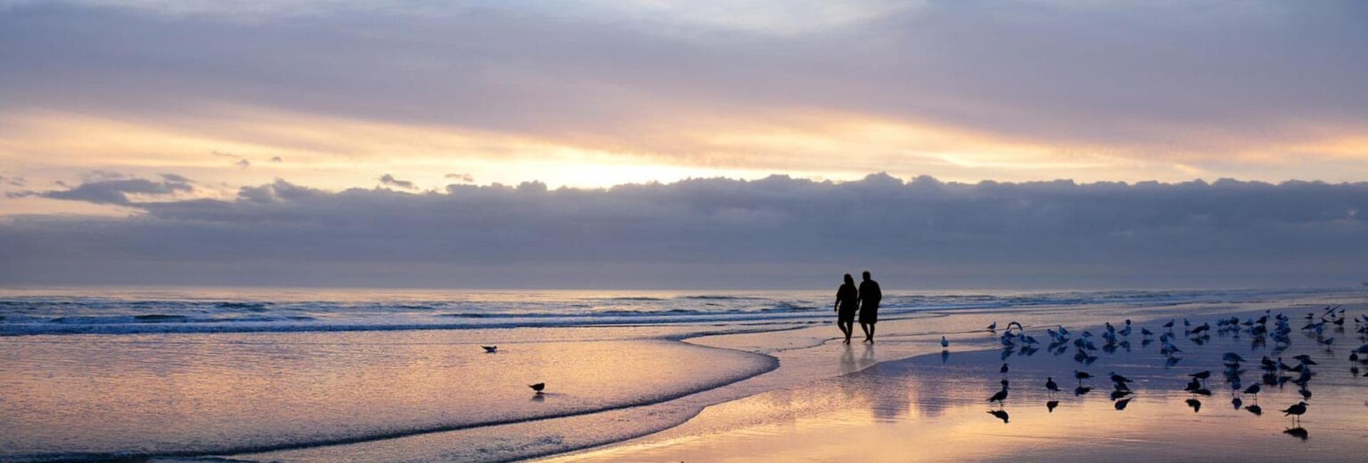 A couple walks hand-in-hand along a sandy beach at sunset, with seagulls scattered nearby.