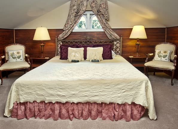King sized bed with antique nightstands and table lamps undre small eyebrow windows with floral curtains. 2 sitting chairs flank the bed and tables.