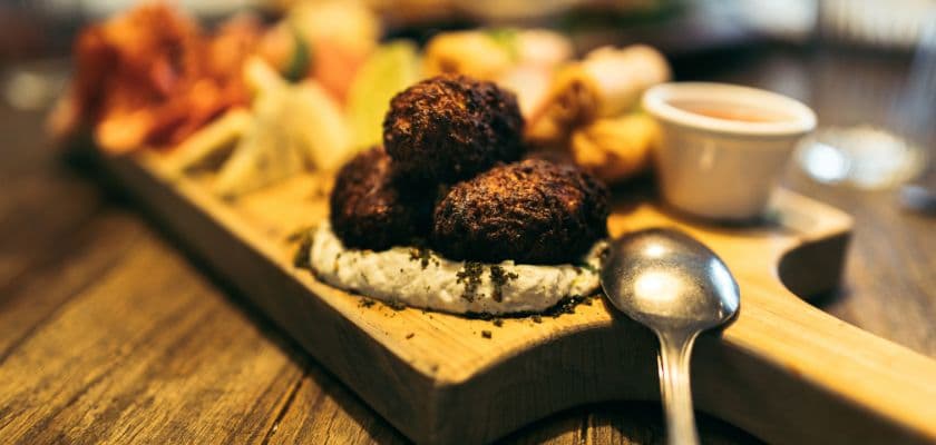 A wooden platter features three golden-brown fritters on a creamy dip, accompanied by various appetizers and a small bowl of sauce.