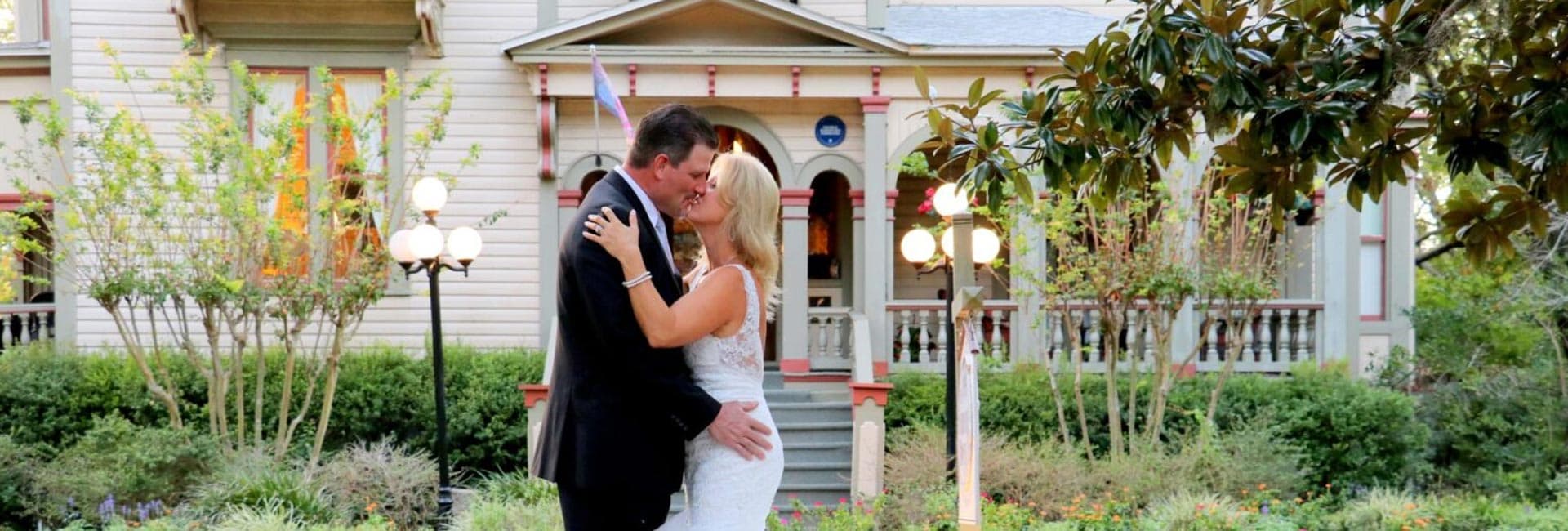 A couple shares a romantic kiss in a landscaped garden in front of a charming house.