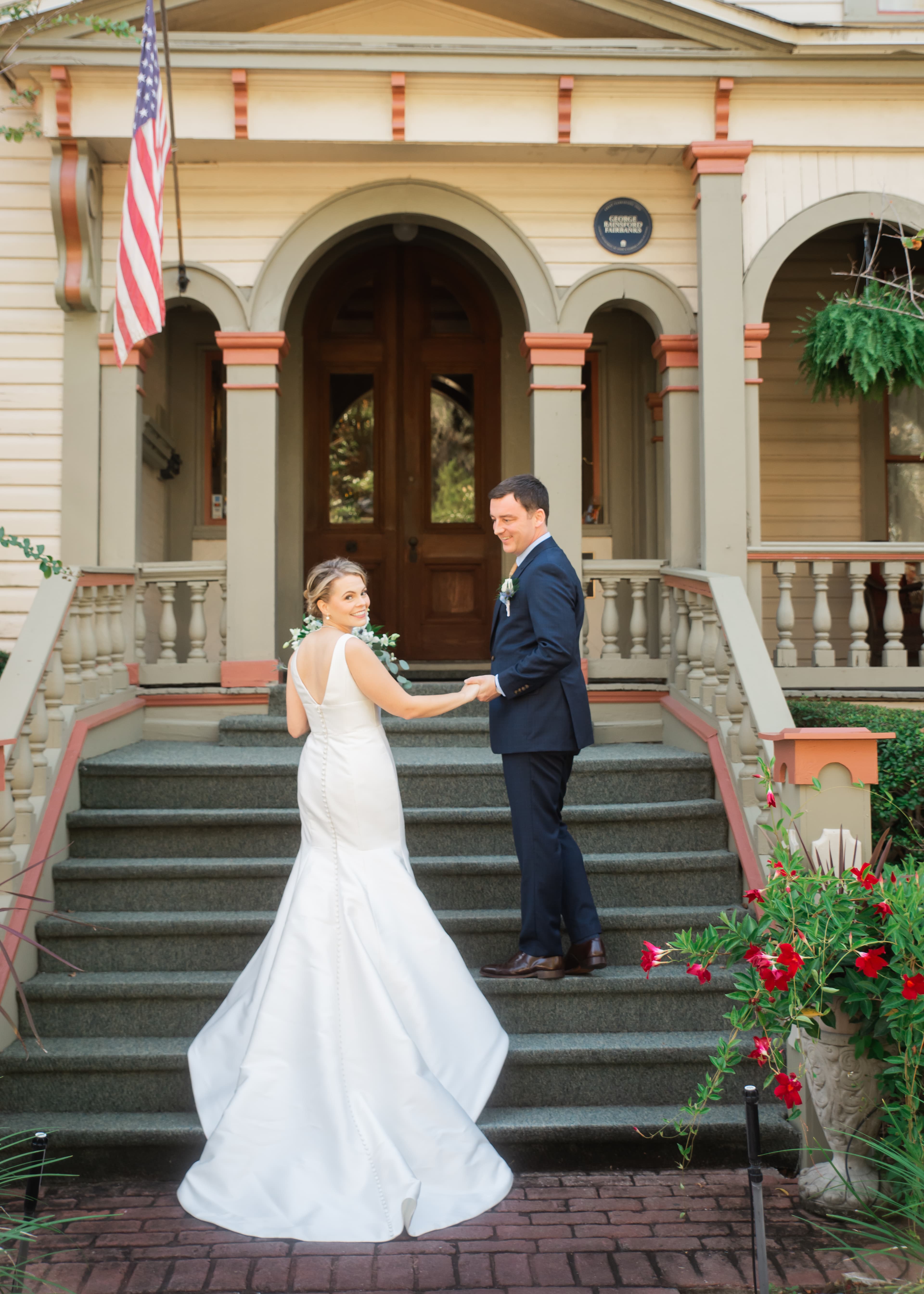 A bride and groom hold hands and smile at each other on the steps of a charming house, adorned with greenery and an American flag.