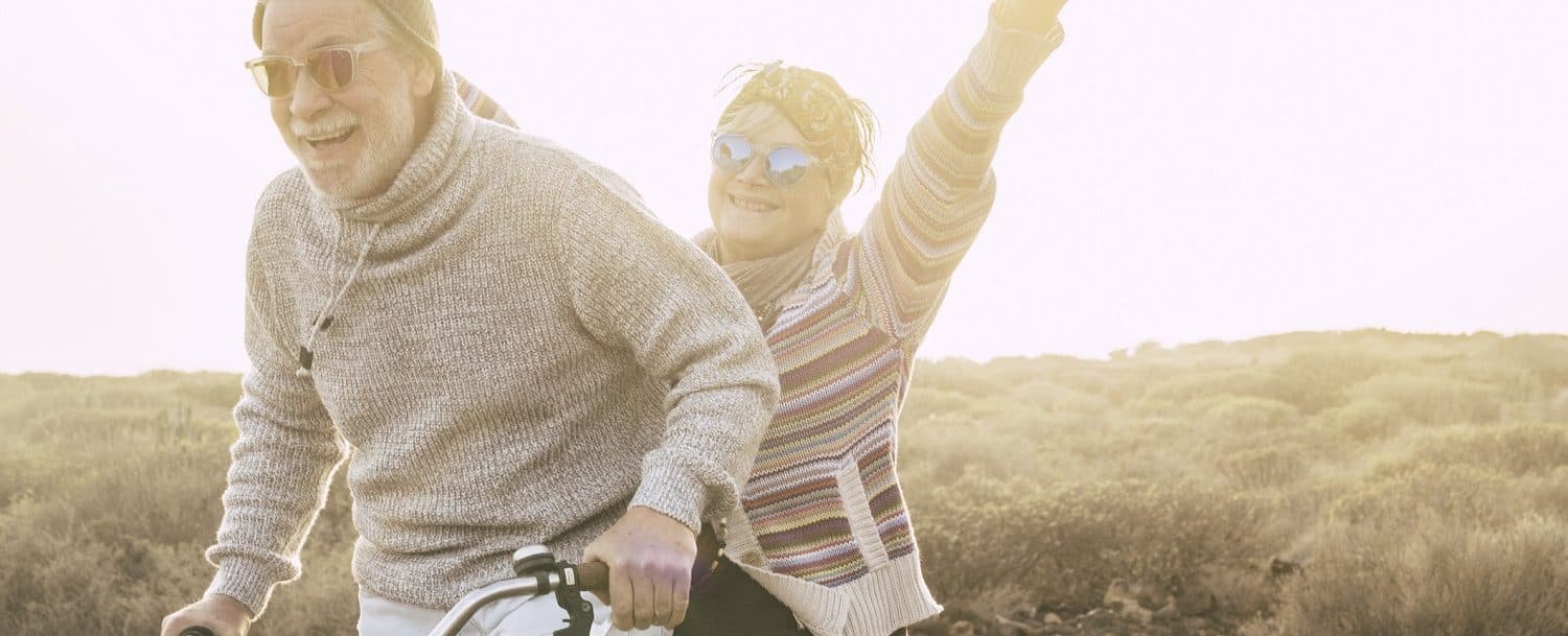 An elderly couple smiles joyfully while riding a bicycle in a sunlit outdoor setting.