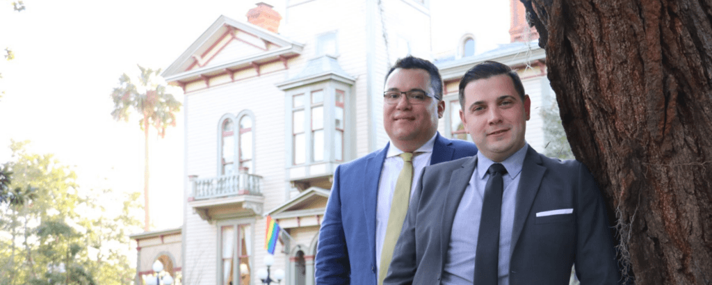 Two men in business attire pose in front of a historic house with palm trees and a rainbow flag.