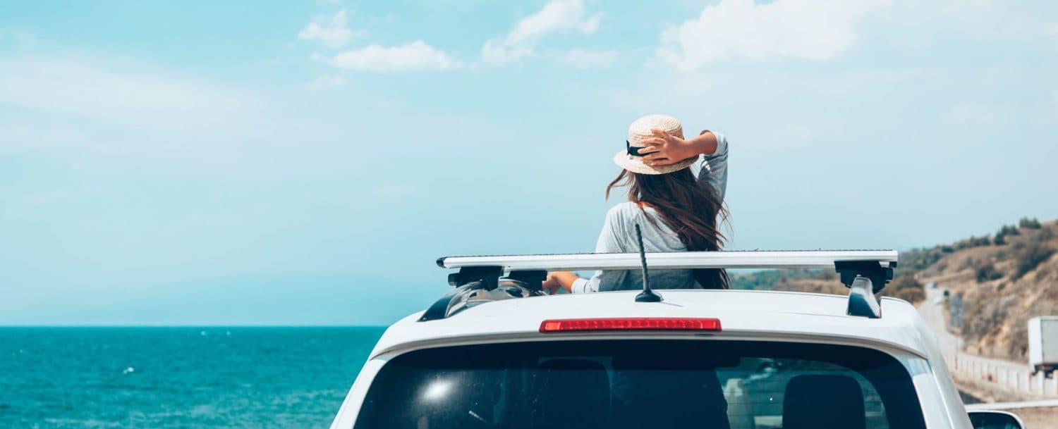 A woman wearing a hat leans back on a car rooftop, looking out at the ocean under a blue sky.