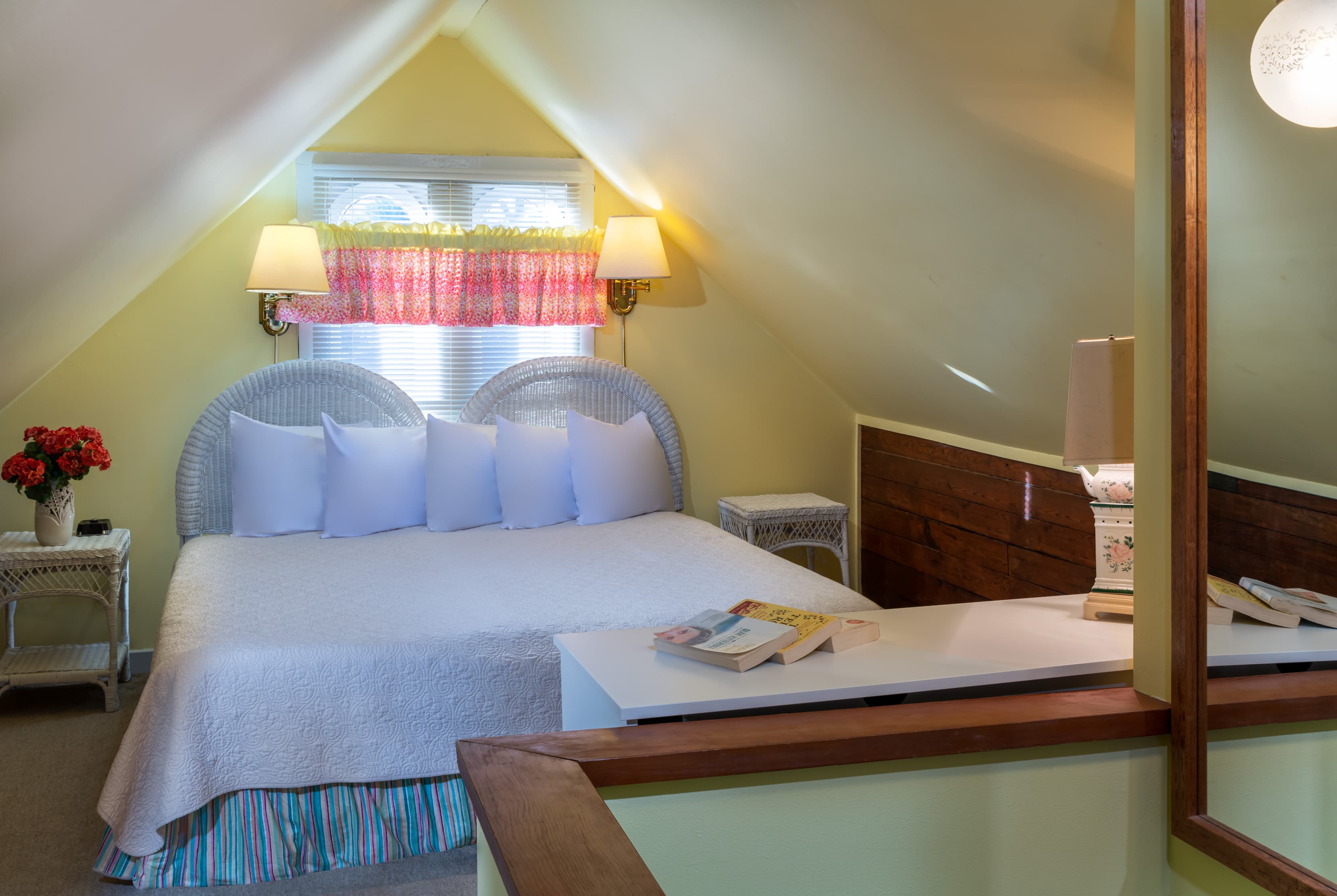 Cozy loft-style bedroom featuring a queen bed with wicker headboard, vaulted ceilings, soft lighting, and colorful cottage accents.