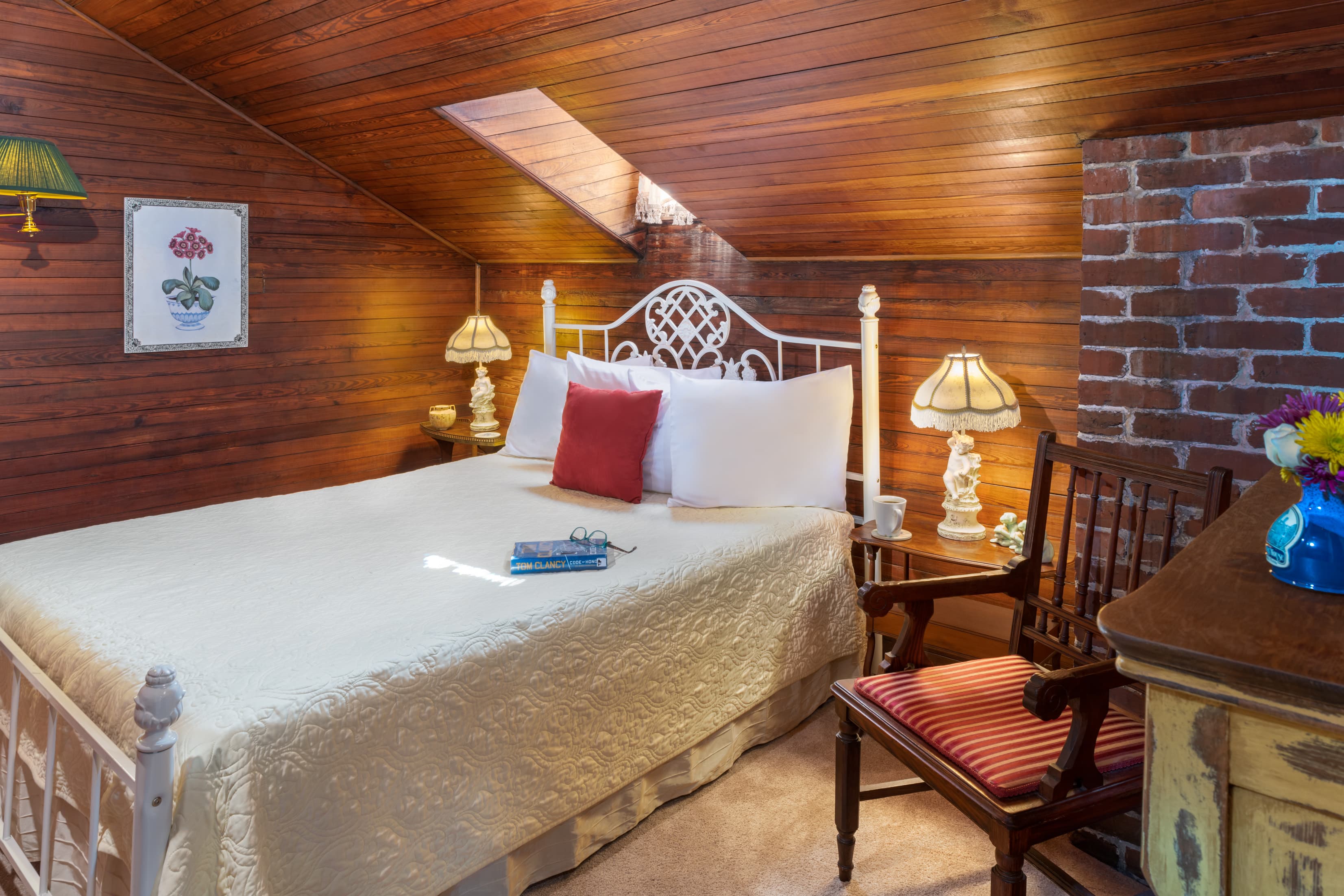 Cozy loft bedroom featuring warm wood-paneled walls, exposed brick accents, vintage lighting, and a comfortable queen bed beneath a skylight.