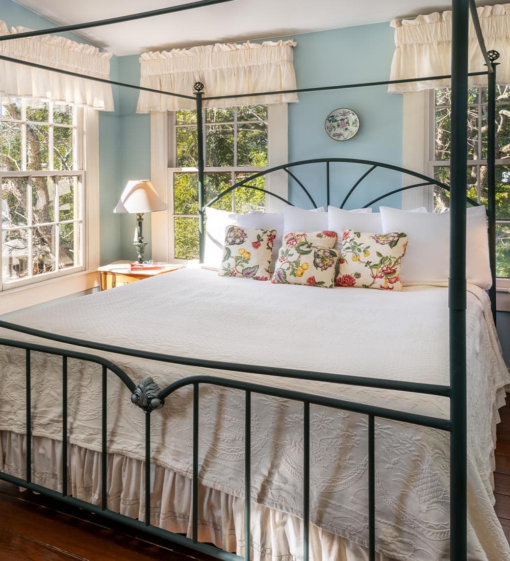 A canopy bed with floral pillows is positioned by windows in a bright, pastel-colored bedroom.