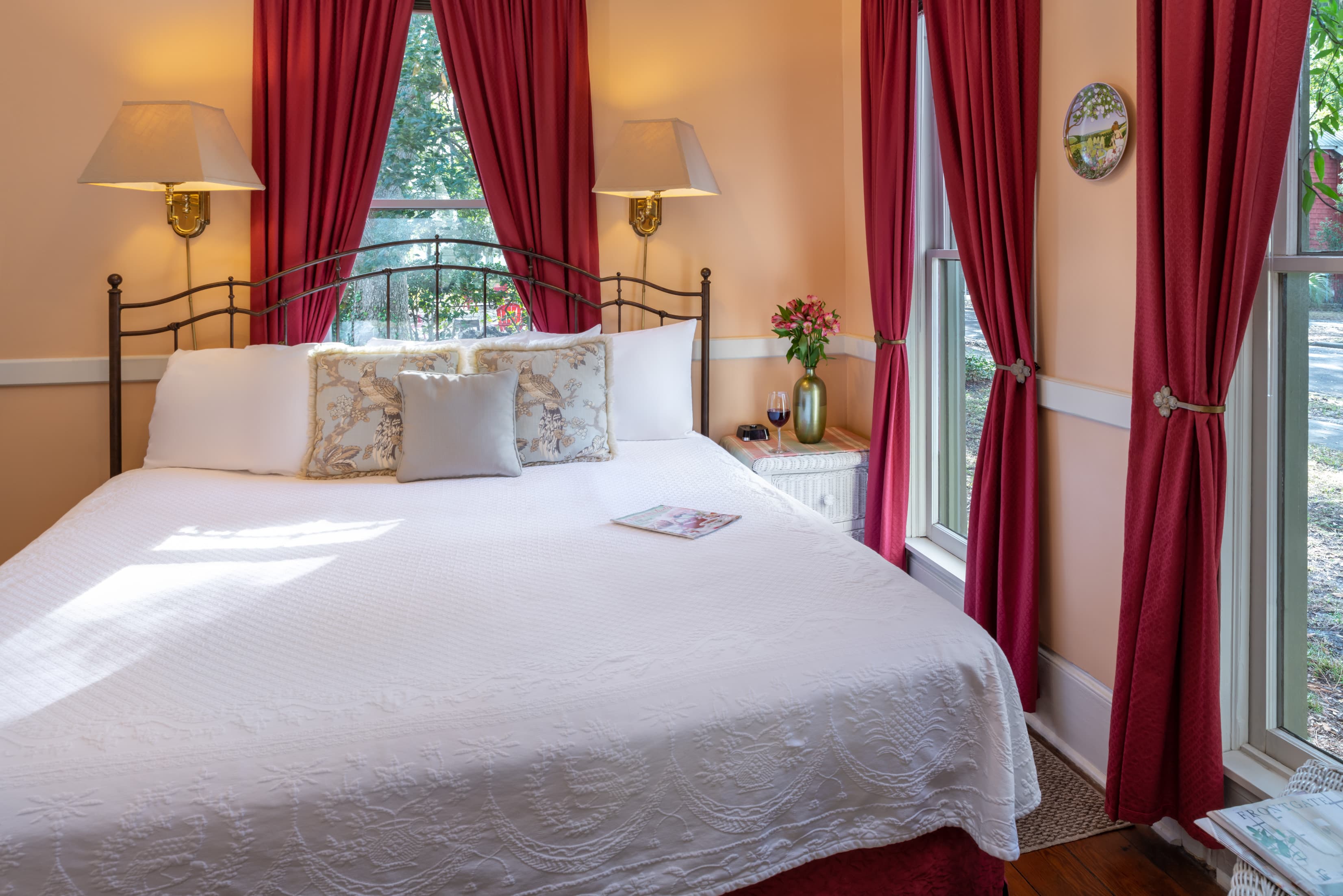 Bright and inviting guest bedroom featuring a king bed with white linens, elegant red drapes, hardwood floors, and large sunlit windows.