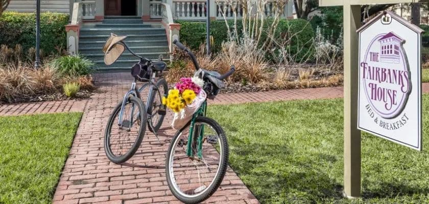 Two bicycles parked on a brick path outside The Fairbanks House Bed and Breakfast, with flowers, green lawn, and the inn sign nearby.