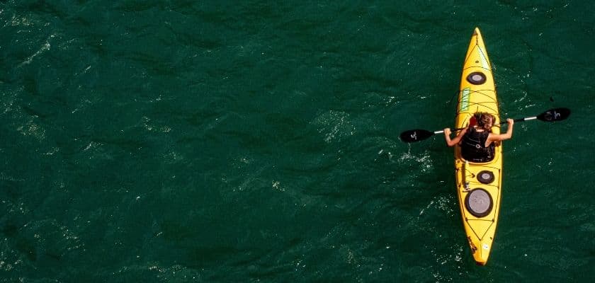 A person kayaking in a bright yellow kayak across deep green water, viewed from above.