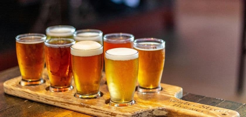A craft beer tasting board with eight small glasses of beer on a wooden paddle.