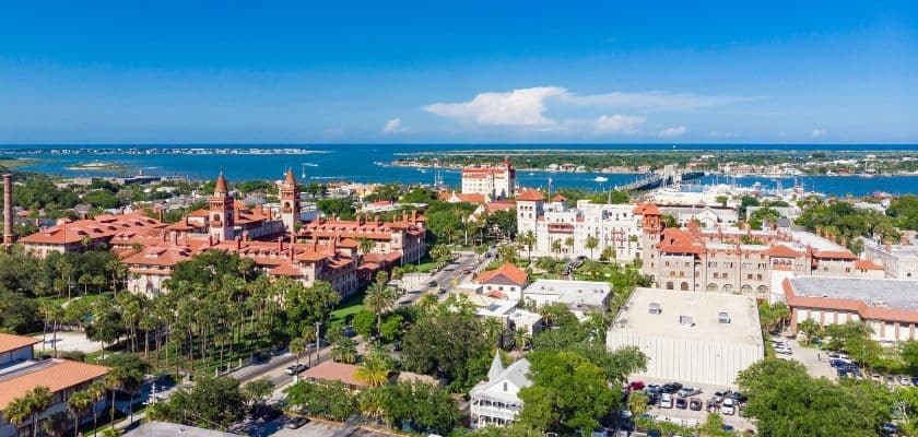 Aerial view of historic St. Augustine, Florida, with red-roofed buildings, palm trees, marinas, and blue water in the distance.