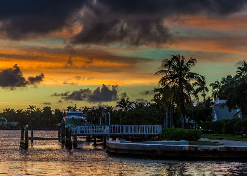 Palm trees and waterfront homes along a dock at sunset with colorful clouds over the water.