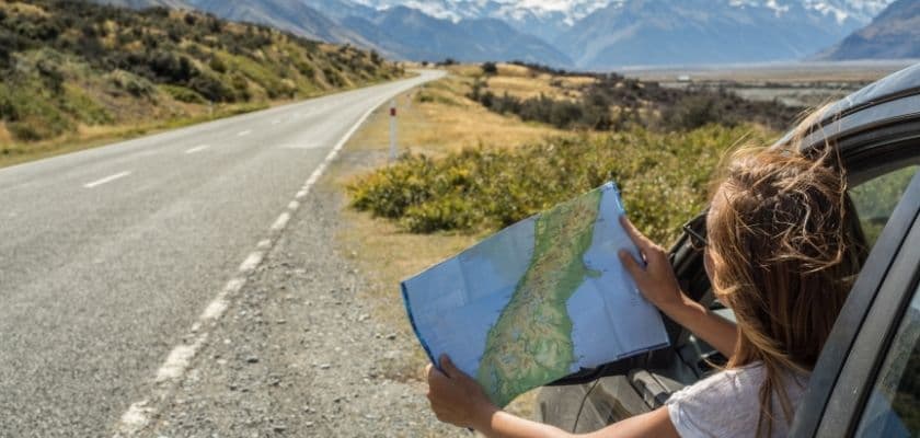 woman leaning out of a car window while holding a map, looking down a winding road surrounded by mountains