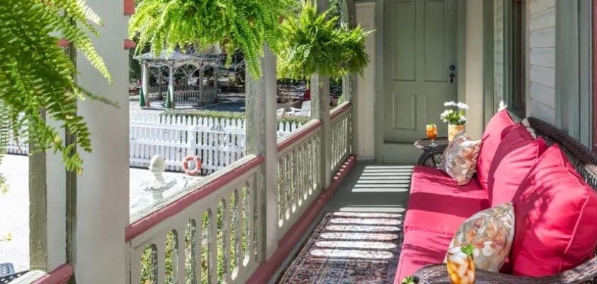 Inviting porch with red cushioned seating and hanging ferns overlooking a white picket fence.