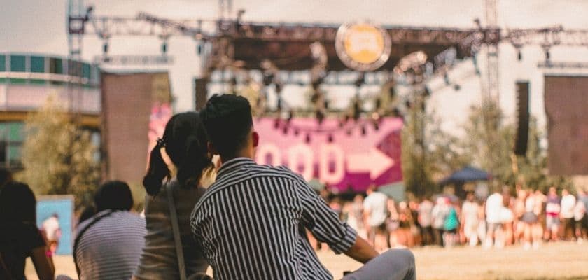 A couple sitting together at an outdoor live performance with a stage and crowd in the background.